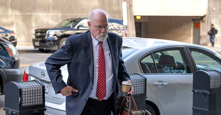 John Durham walking through a parking garage carrying a coffee and a bag