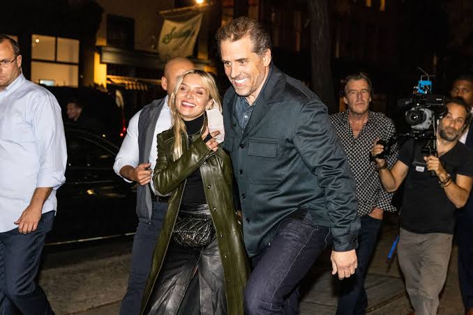 Hunter Biden walking arm in arm with a companion on a New York street at night, surrounded by photographers