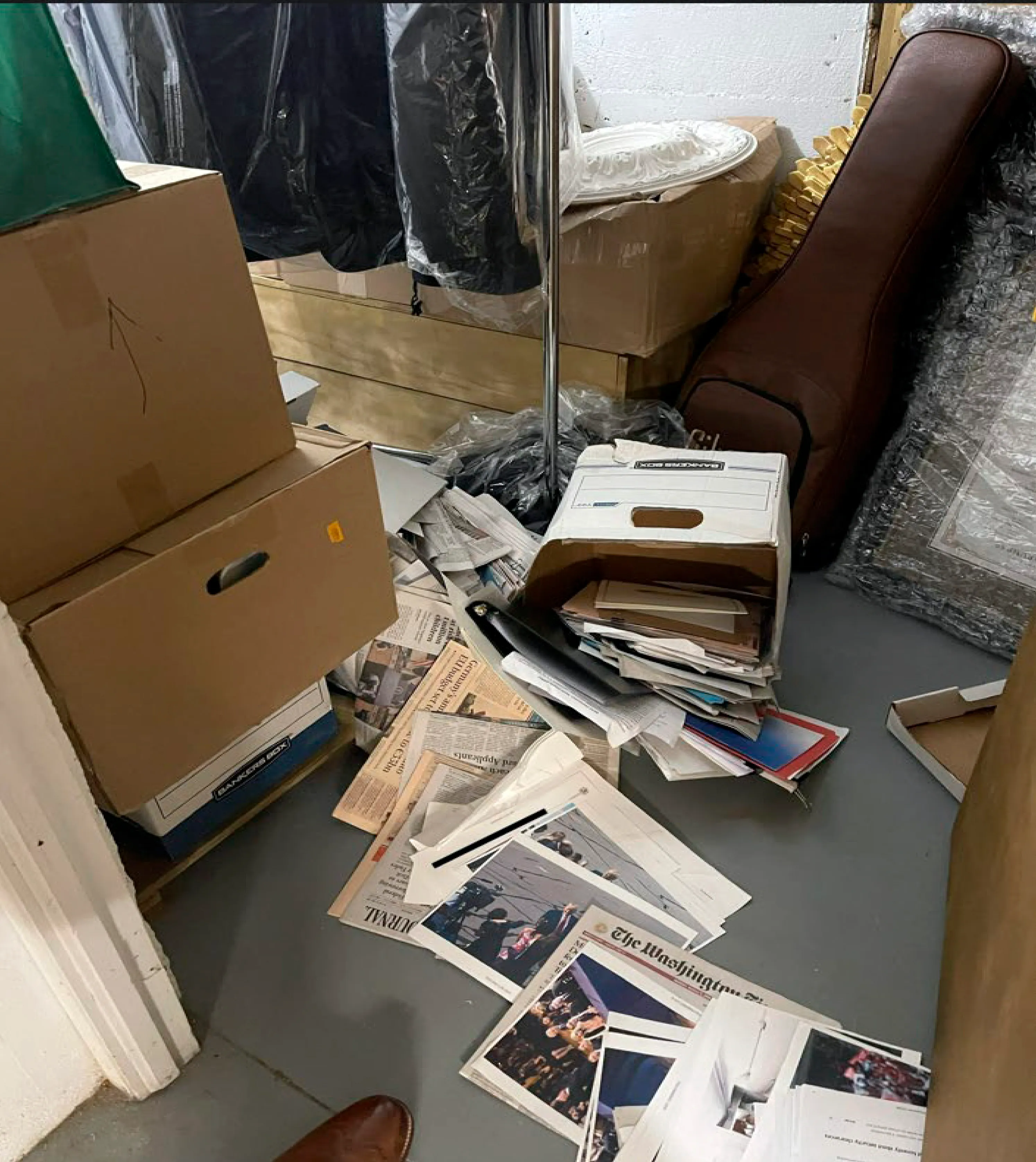 A storage room with banker's boxes torn open and newspapers, magazines, and loose photographs spilled across the floor
