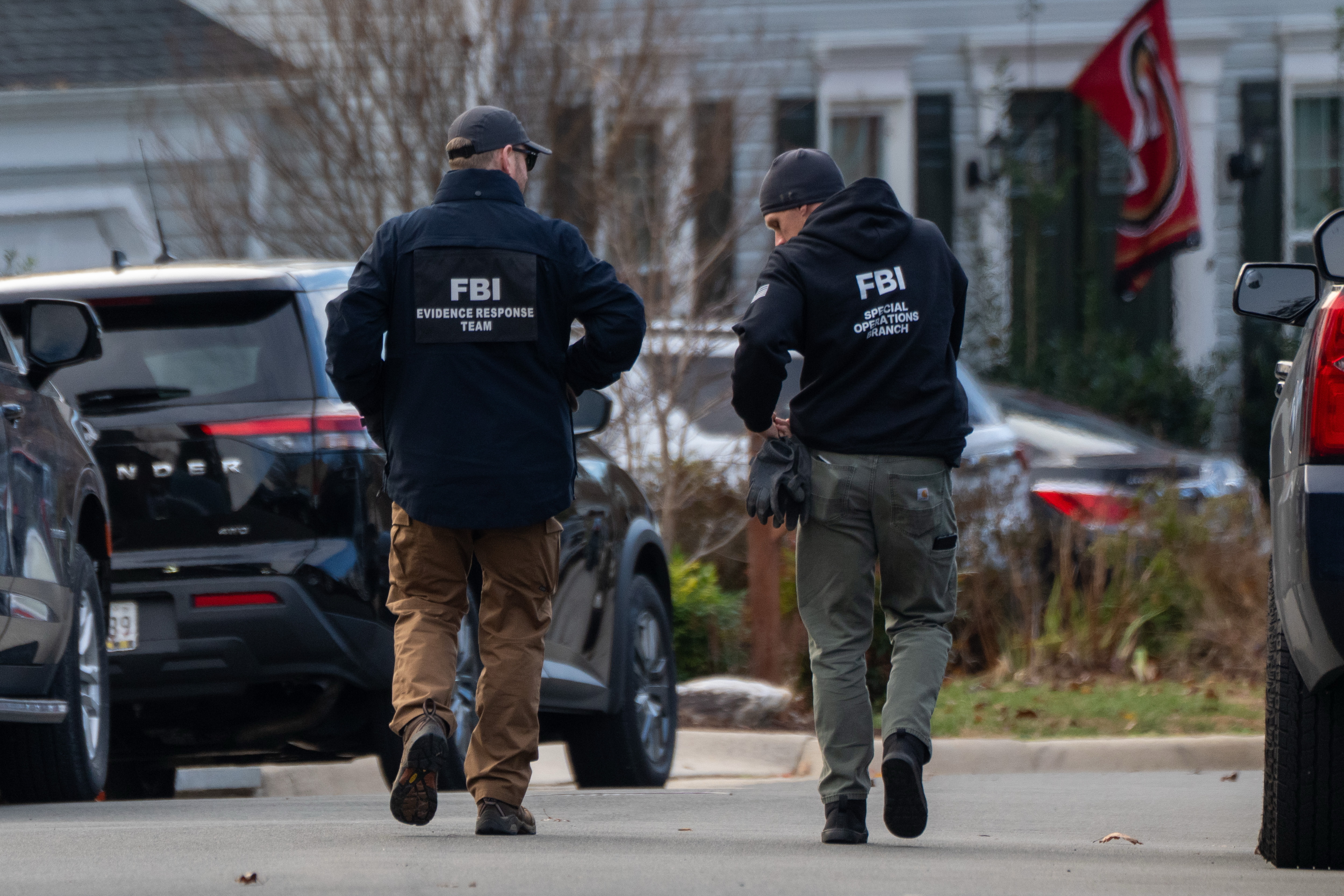 Two FBI agents seen from behind walking down a residential street lined with parked SUVs, one in a jacket marked Evidence Response Team and the other in a hoodie marked Special Operations Branch pulling on gloves, with a white house and a red flag in the background