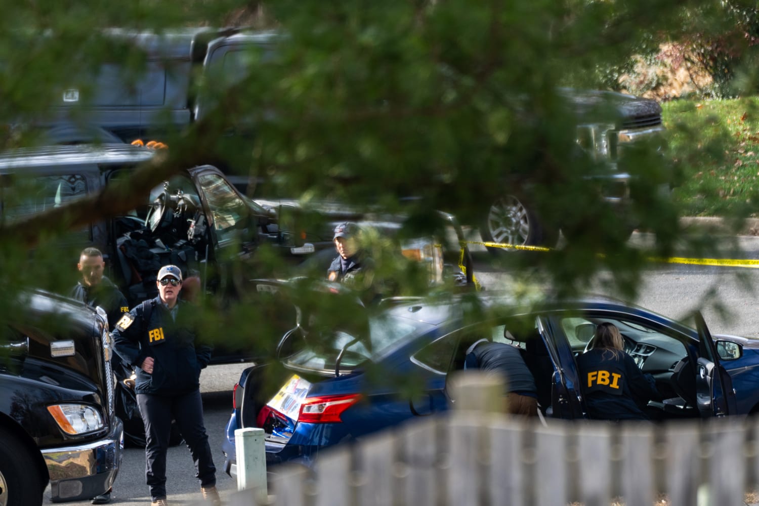 Seen through tree branches and over a wooden fence, FBI agents in marked jackets search a blue sedan with its trunk and both doors open, one agent leaning into the driver's seat, beside parked SUVs and a line of yellow police tape