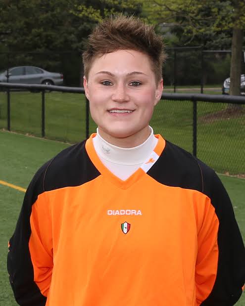 A smiling young person with short spiky brown hair in an orange and black Diadora goalkeeper jersey over a white turtleneck, standing on an artificial turf soccer field in front of a black chain-link fence and trees
