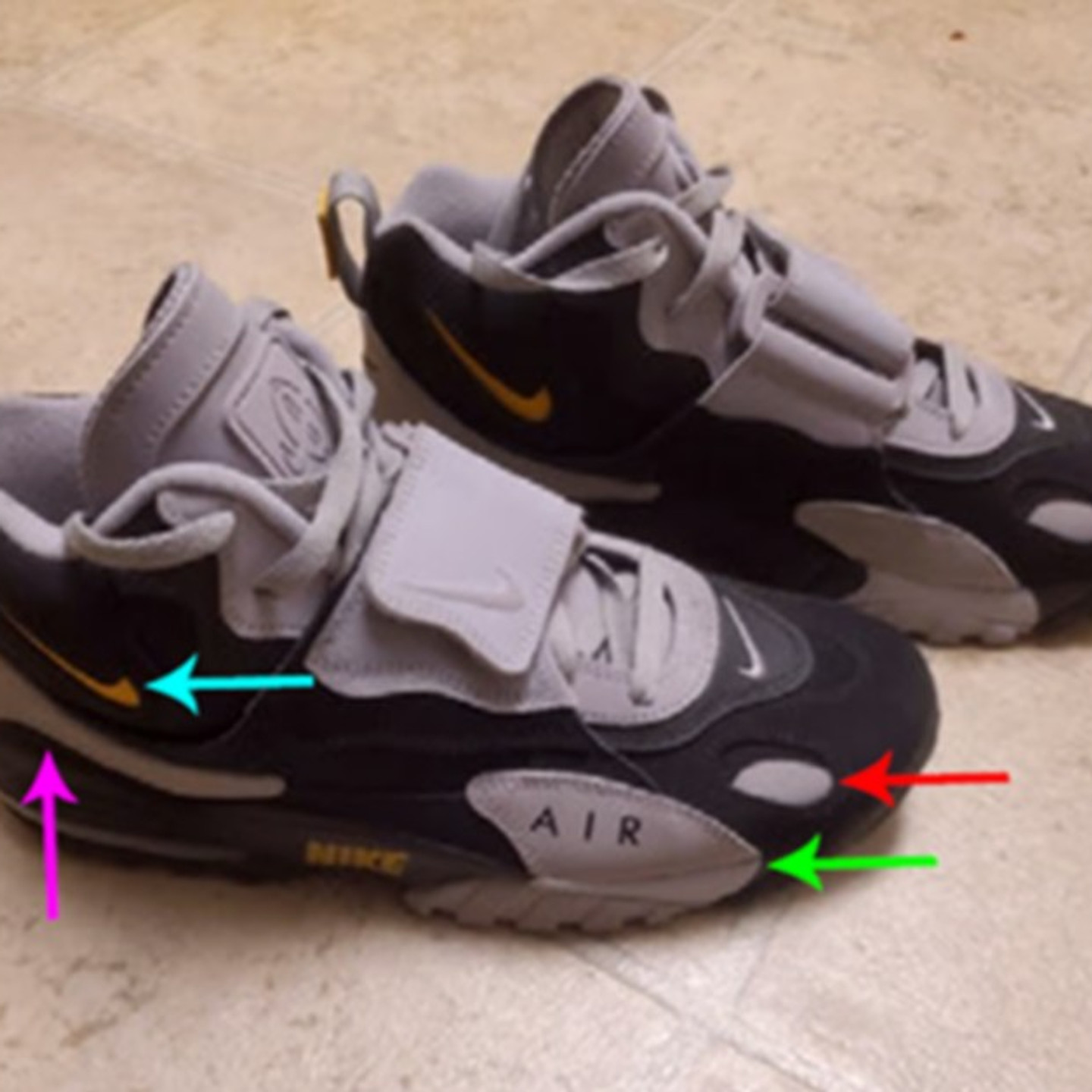 A pair of black and gray Nike Air Max Speed Turf sneakers on a tile floor, with colored arrows pointing to the yellow swoosh on the side, the light stripe along the heel, the oval cutout near the toe and the gray AIR panel on the forefoot