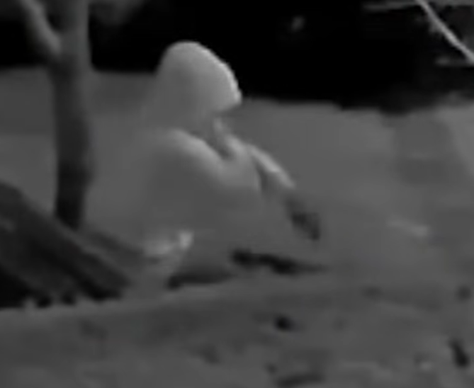 An extremely blurry, enlarged grayscale crop of surveillance footage showing the hooded suspect from the side, the hood and shoulders visible as soft gray shapes with no discernible facial features