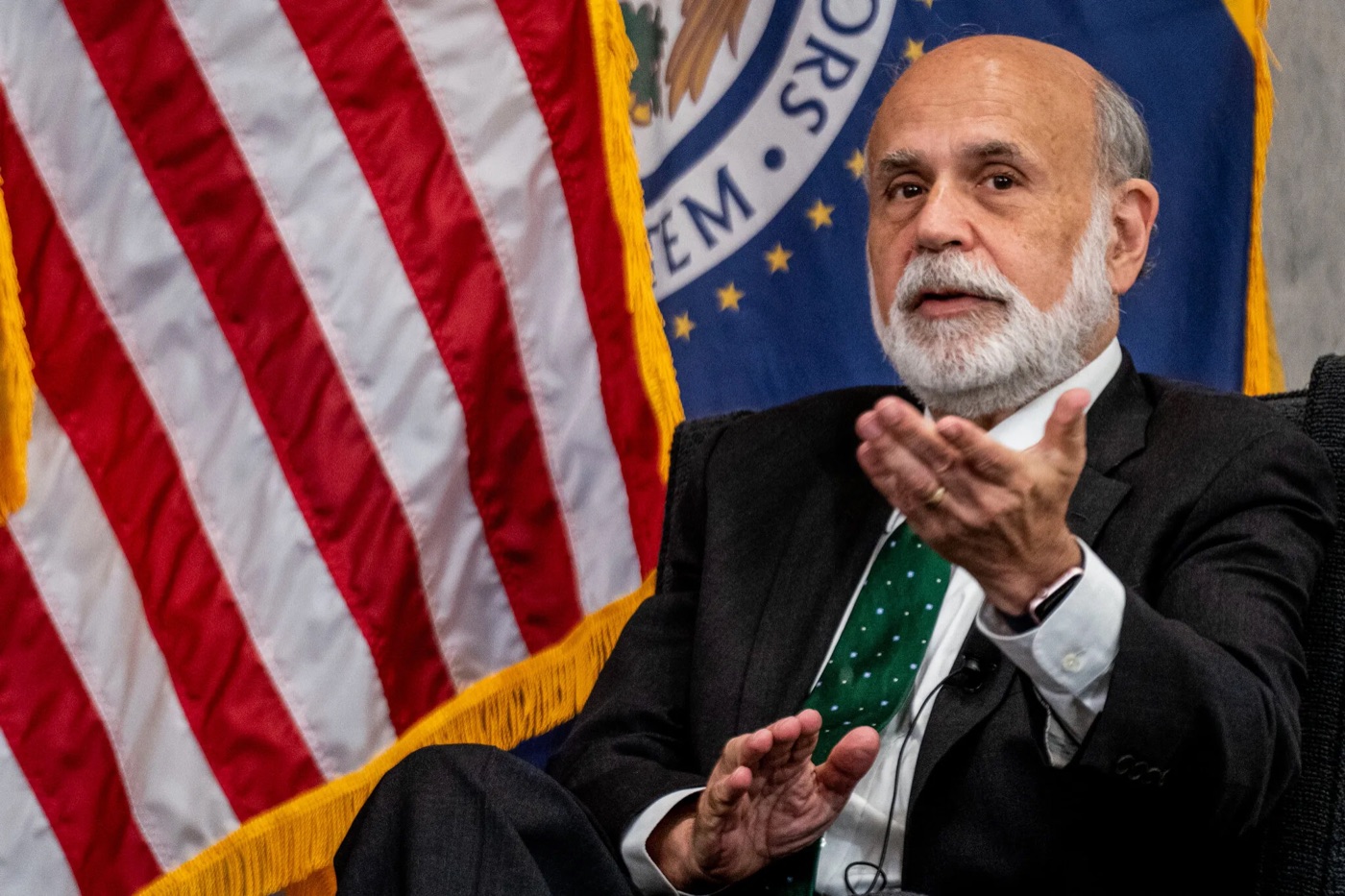 Ben Bernanke, gray-bearded, speaking and gesturing with both hands while seated in front of an American flag and the flag of the Federal Reserve Board of Governors
