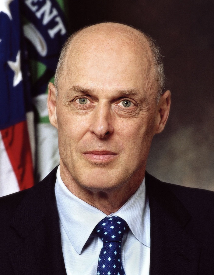 Official 2006 Treasury Department portrait of Henry Paulson in a dark suit and blue patterned tie, with the American and Treasury flags behind him