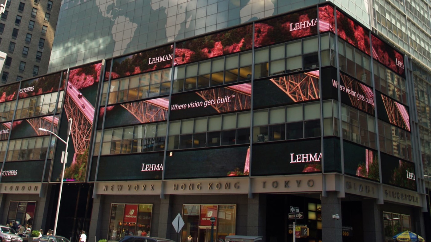 Lehman Brothers headquarters at 745 Seventh Avenue near Times Square, its facade wrapped in giant video screens showing the Lehman name and the slogan Where vision gets built, above a stone band engraved with London, New York, Hong Kong, Tokyo, Miami, and Singapore