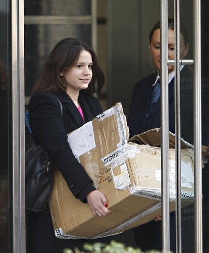 A young Lehman Brothers employee in a black coat leaving through the building's glass doors carrying a battered, taped-up cardboard box, as a security guard holds the door behind her