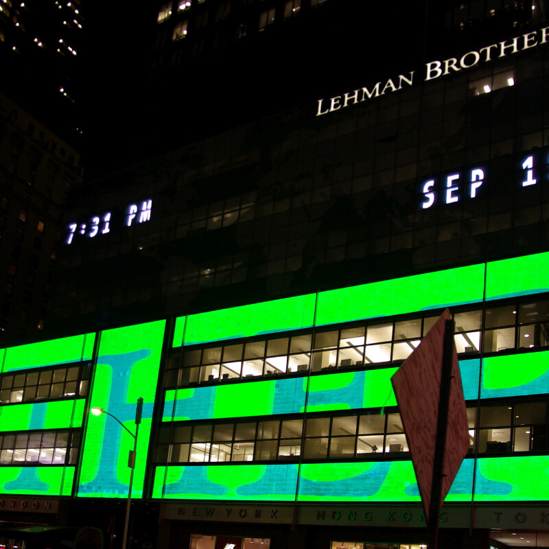 Lehman Brothers headquarters at night, the name lit in white letters near the top of the tower beside a display reading 7:31 PM and SEP 15, above floors of video screens glowing solid green