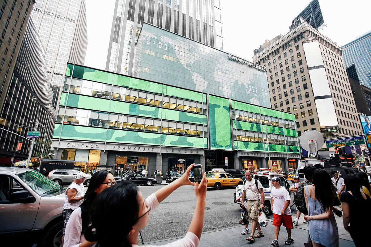 Tourists on the sidewalk photographing Lehman Brothers headquarters near Times Square with their phones, the building's wraparound screens glowing bright green with giant Lehman lettering above a stone band reading Chicago, Frankfurt, London, as a television satellite truck sits parked at the curb