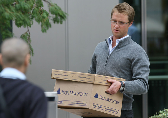 A Lehman Brothers employee in a gray sweater and glasses carrying two stacked Iron Mountain storage boxes out of the firm's London offices