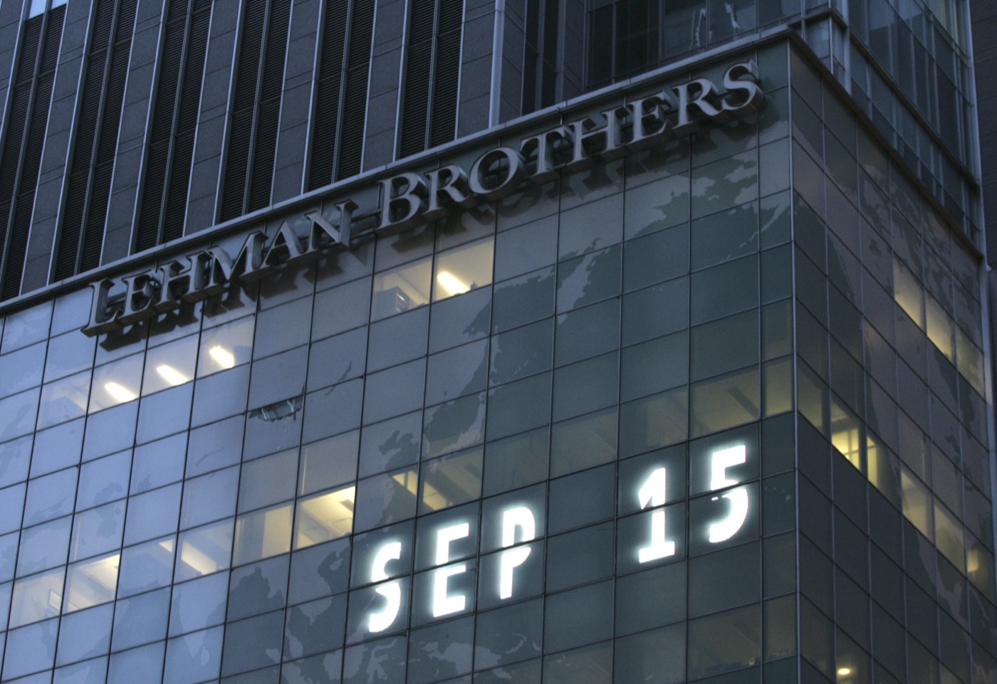 The Lehman Brothers name in raised steel letters on the headquarters facade at dusk, with the building's lit ticker display reading SEP 15 across the glass below it