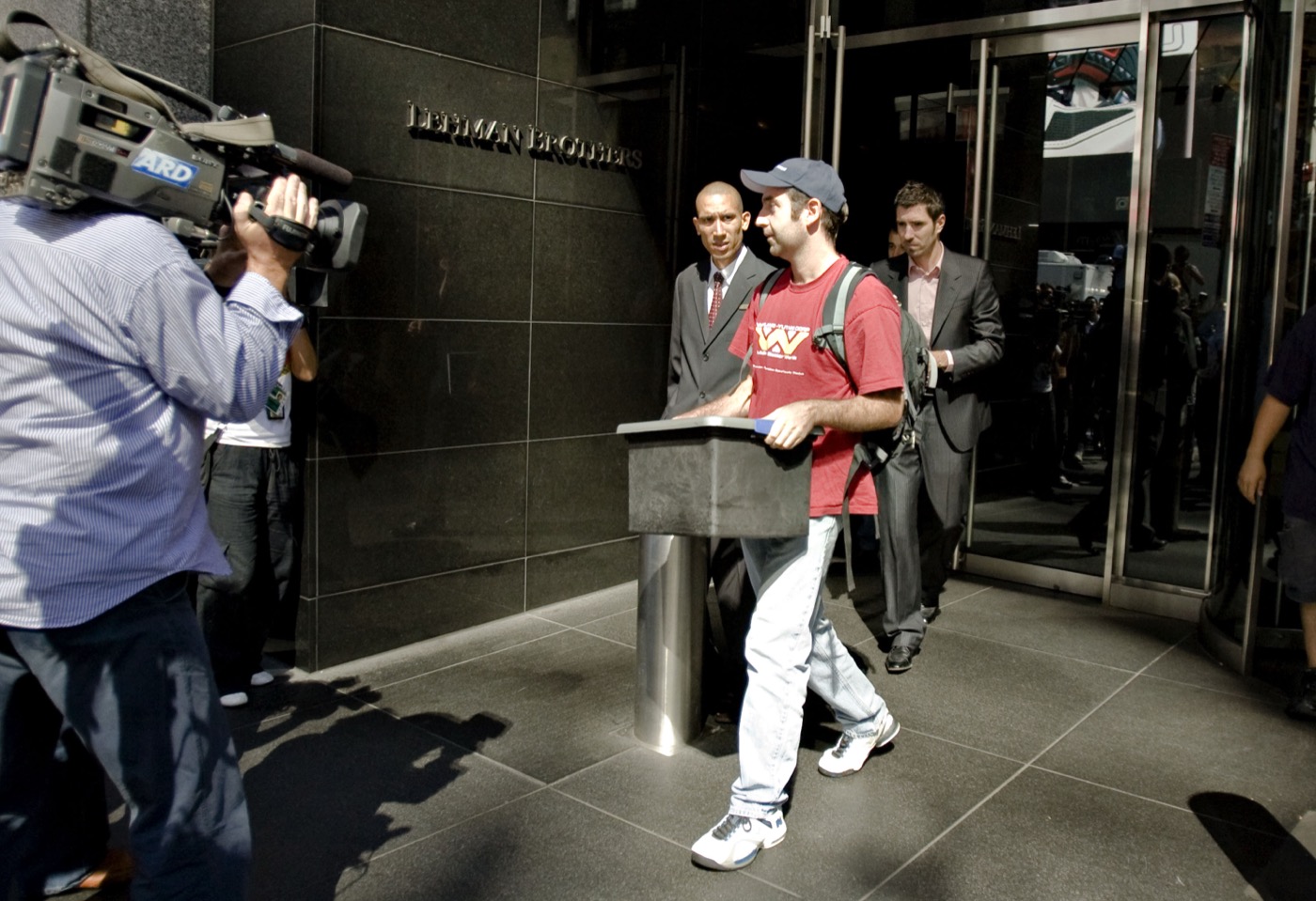 A Lehman Brothers employee in a red t-shirt and baseball cap carrying a plastic storage bin out of the firm's headquarters at 745 Seventh Avenue, past the Lehman Brothers sign on the granite facade, as a German ARD television cameraman films him and a security guard looks on