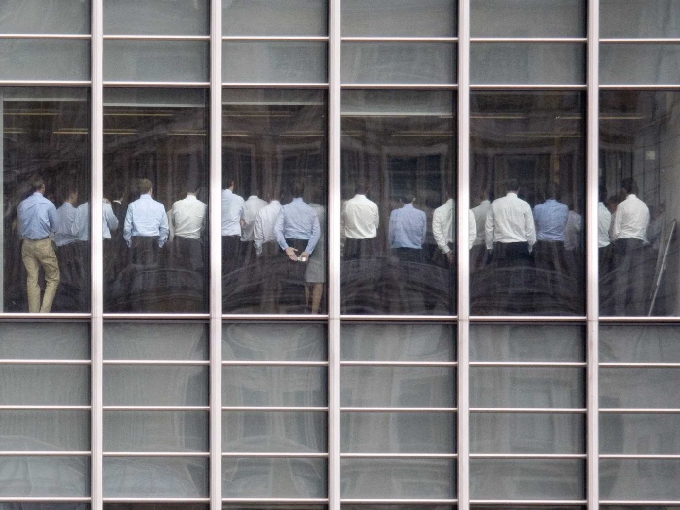Seen from outside through a grid of office windows, a long row of Lehman Brothers employees in shirtsleeves stand with their backs to the camera, gathered together inside the firm's offices