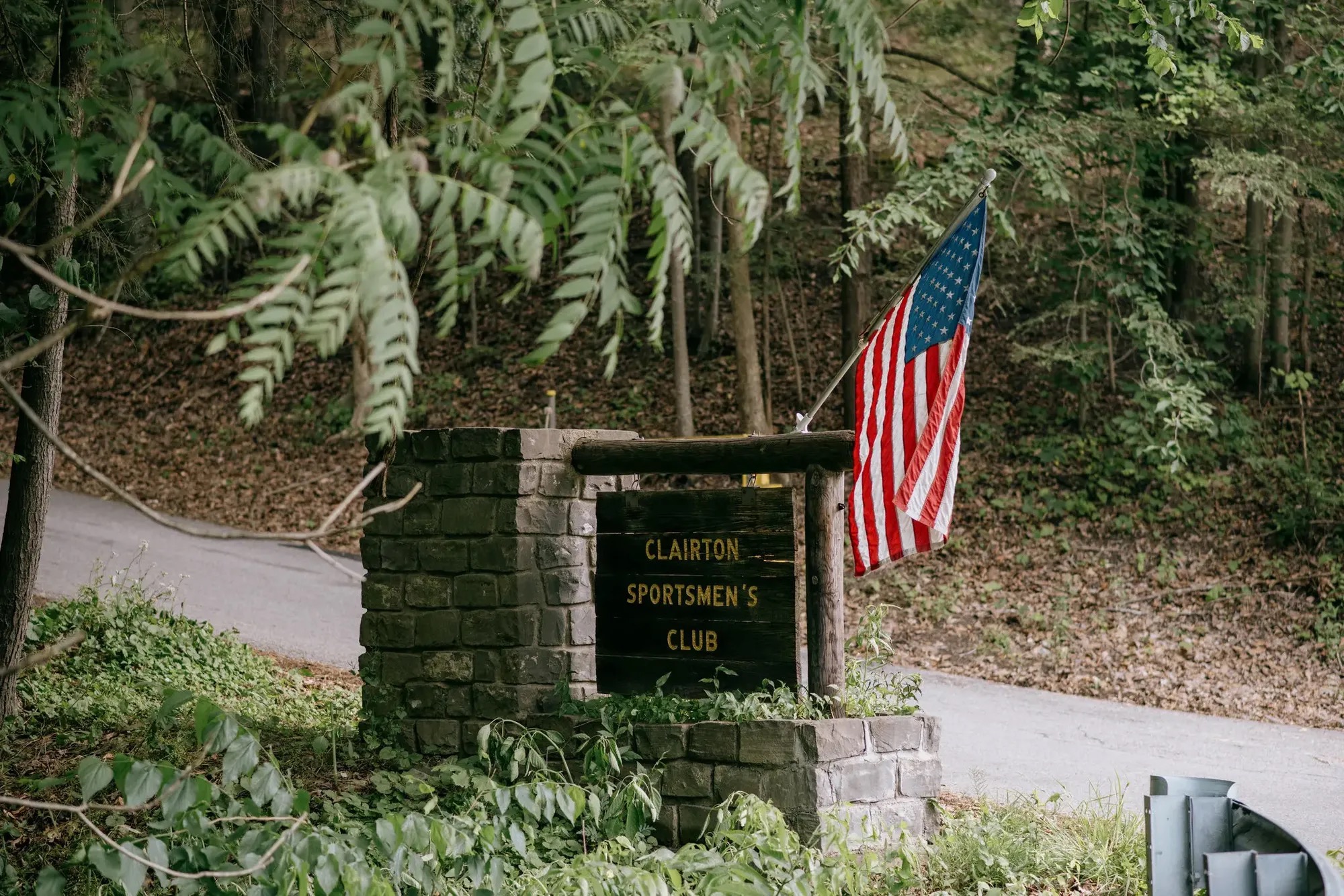 The wooden entrance sign for the Clairton Sportsmen's Club, set into a stone pillar in the woods, with an American flag mounted above it