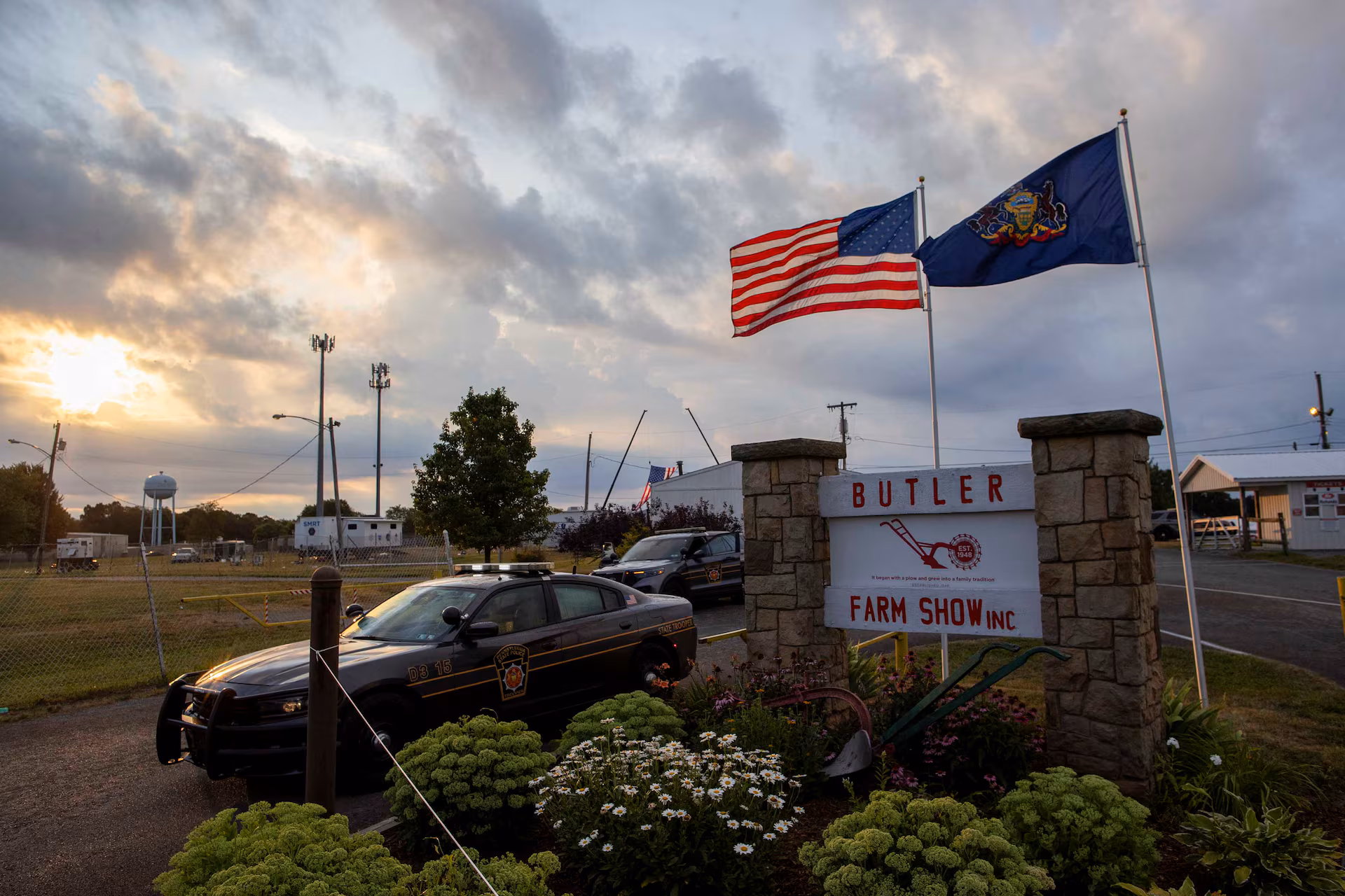 The stone entrance sign for the Butler Farm Show, with police vehicles parked nearby and American and Pennsylvania flags flying above it at dusk