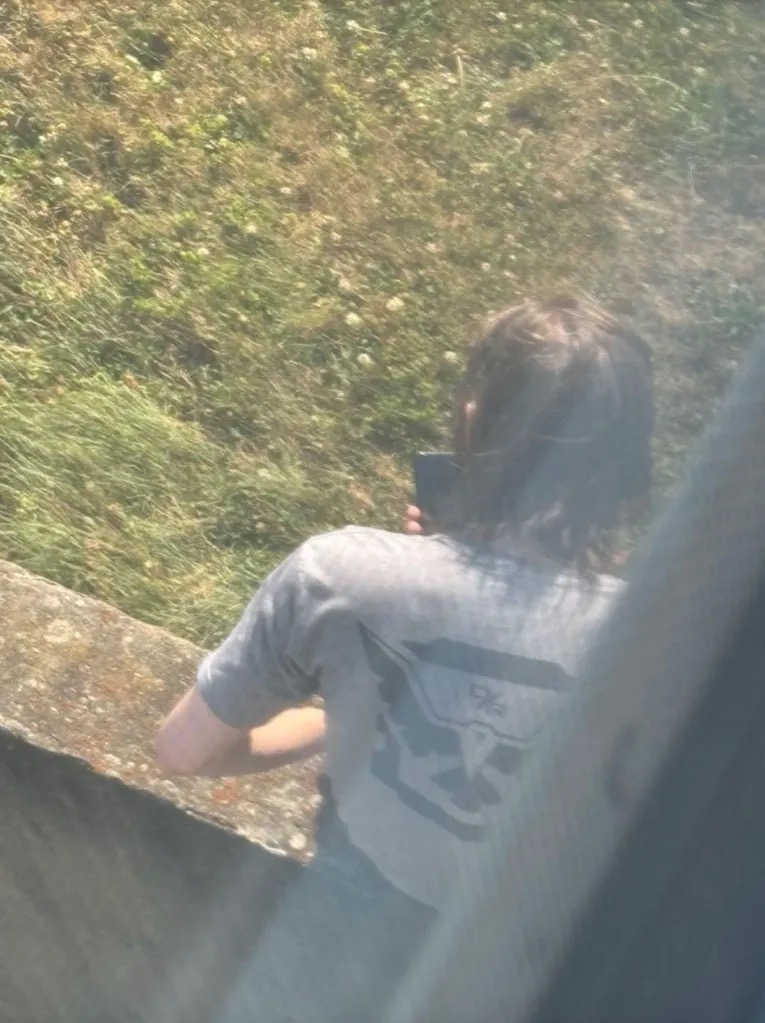 Photograph of Thomas Crooks from behind on the AGR International rooftop shortly before the shooting, wearing a gray t-shirt and holding up a phone