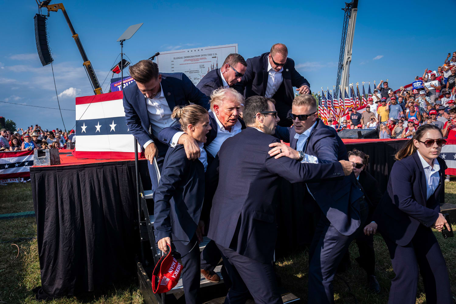 Photograph of Secret Service agents surrounding Donald Trump and rushing him down the stage steps immediately after the shooting, blood visible near his ear, with the crowd and American flags in the background