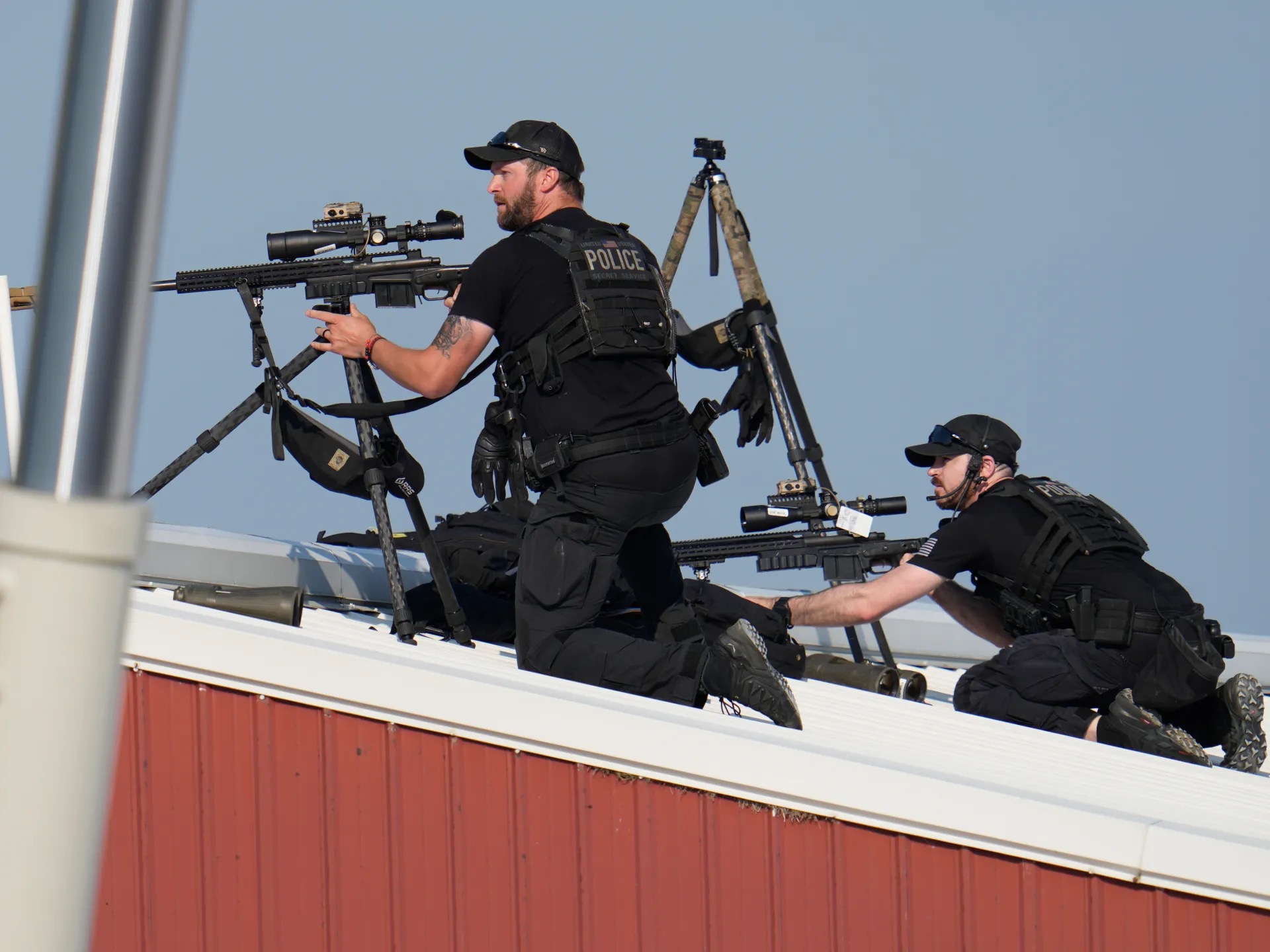 Two Secret Service counter snipers in black tactical gear positioned on a rooftop, one aiming a rifle mounted on a tripod while the other watches through his own scope