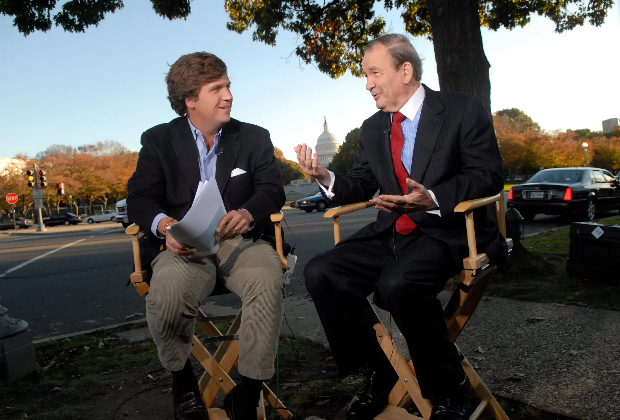 Tucker Carlson interviewing a guest in director's chairs outdoors near the U.S. Capitol