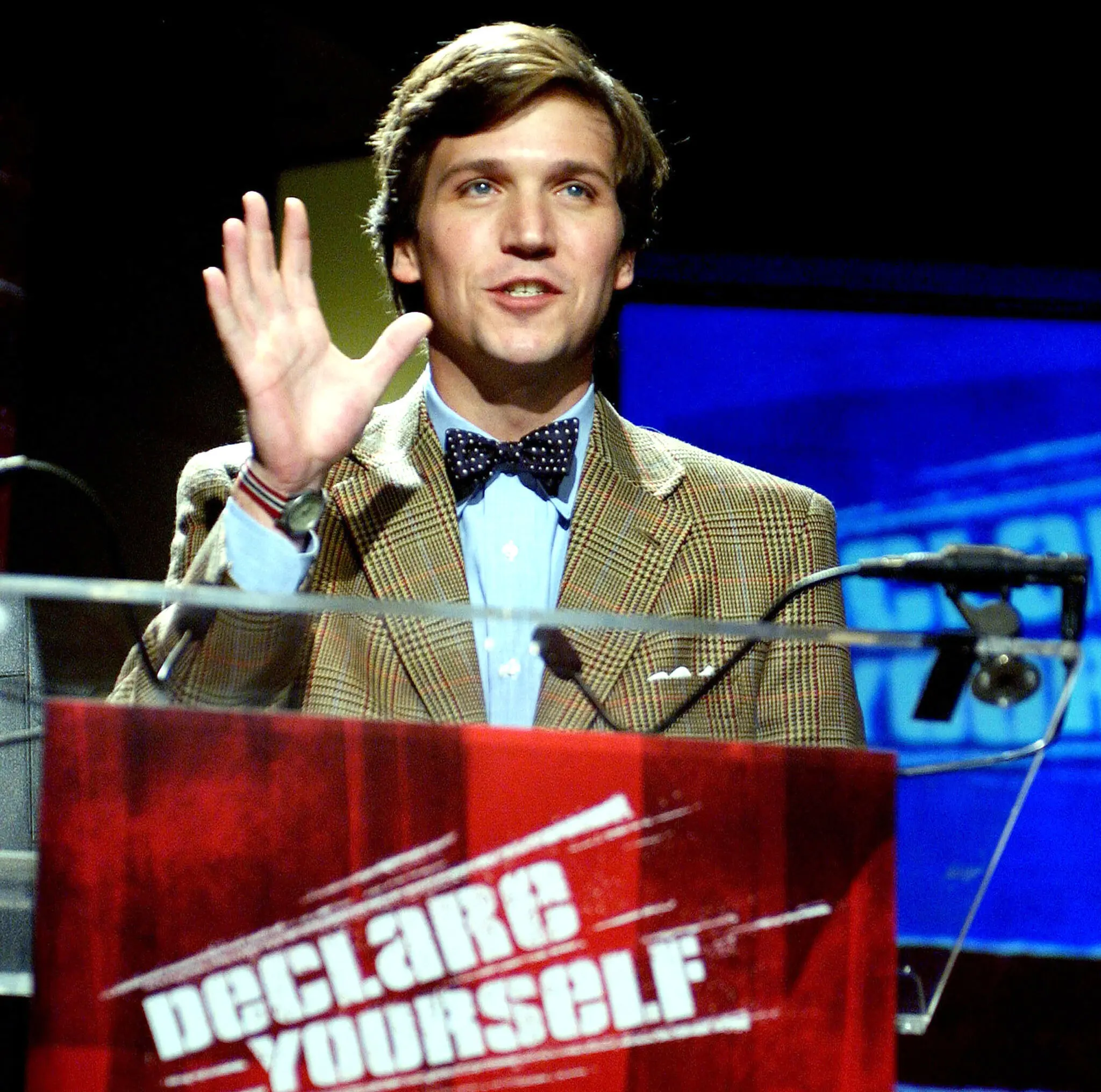 A younger Tucker Carlson in a bow tie and tweed jacket, waving from behind a podium reading Declare Yourself