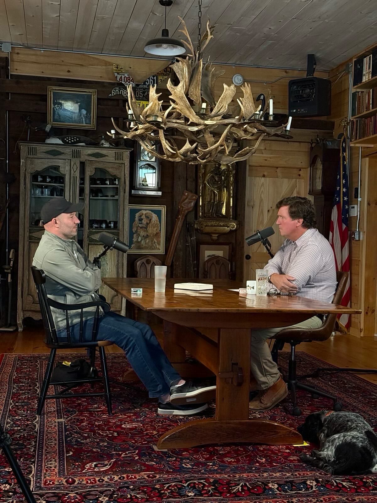 Tucker Carlson interviewing a guest at a wooden table in his own studio in Maine, with an antler chandelier and an American flag