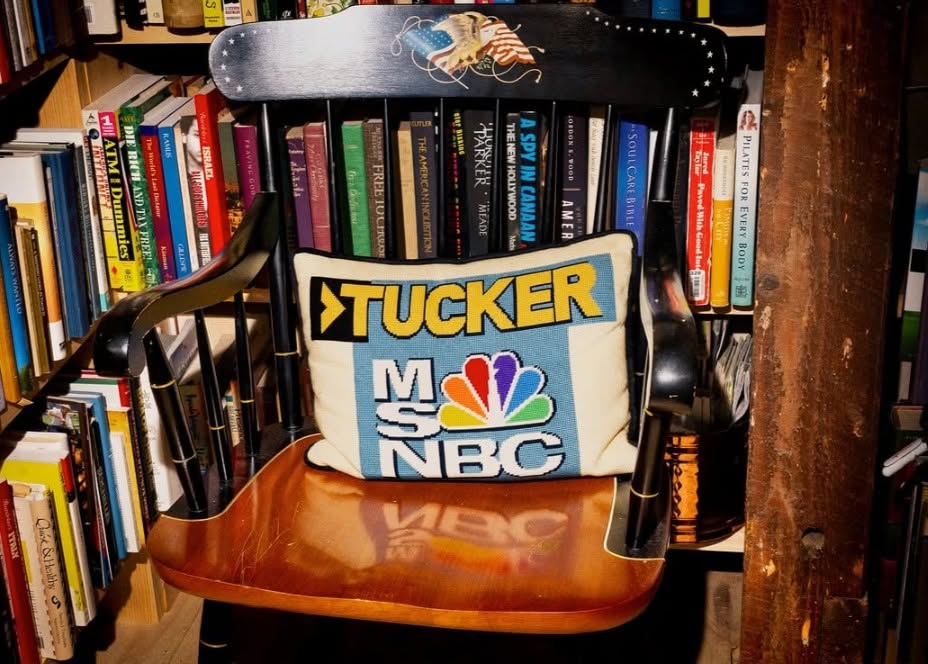 A needlepoint pillow reading TUCKER with the MSNBC logo underneath, on a rocking chair in front of a bookshelf
