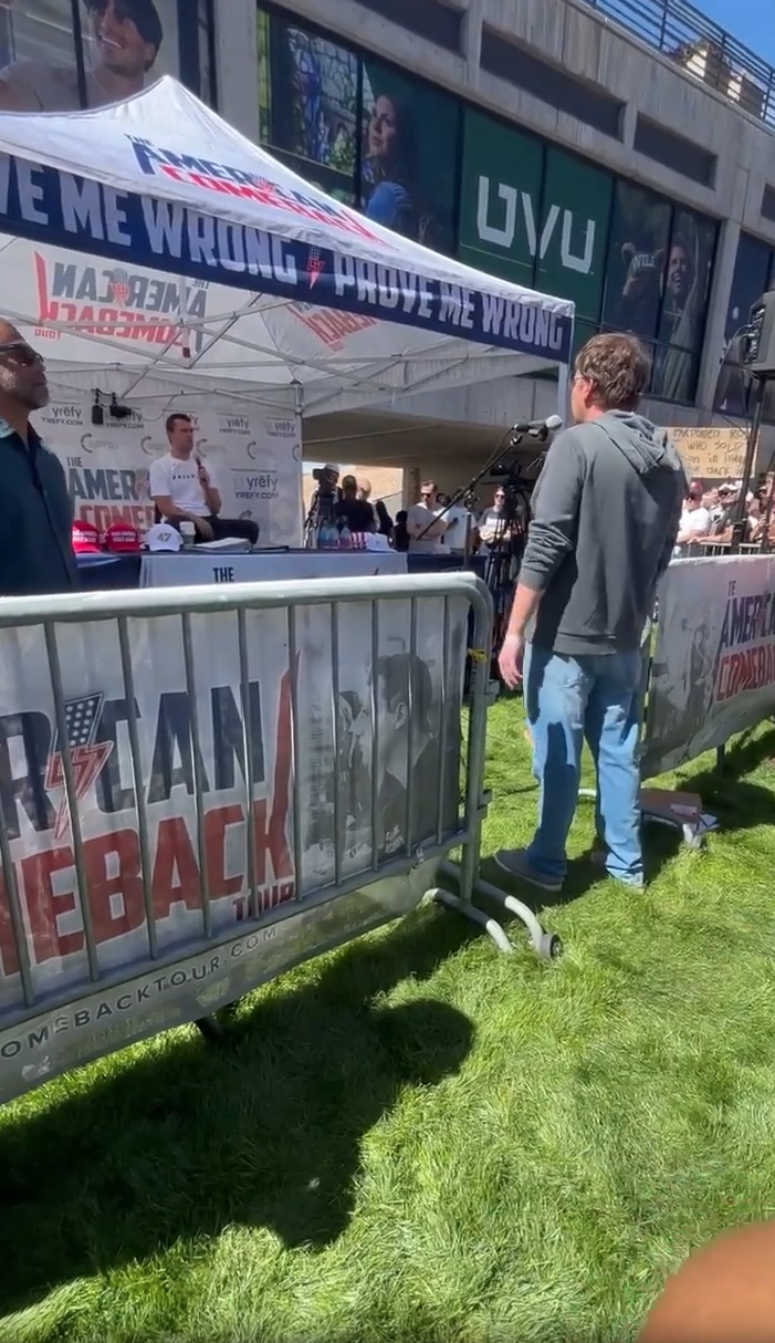 A man in a gray hoodie standing near the metal barricade separating the crowd from the tented stage area, with the UVU building visible in the background