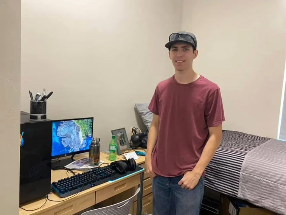 Photo of Tyler Robinson in his Utah State University dorm room in fall 2021, standing beside a desk with a gaming keyboard, headset, and monitor