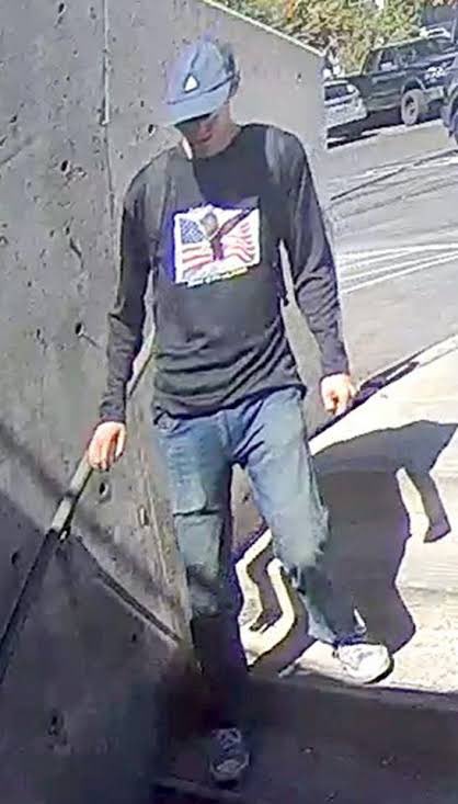FBI person-of-interest photo showing a man in a black long-sleeve shirt printed with an American flag and eagle, cap, and sunglasses, walking on a ramp near Utah Valley University