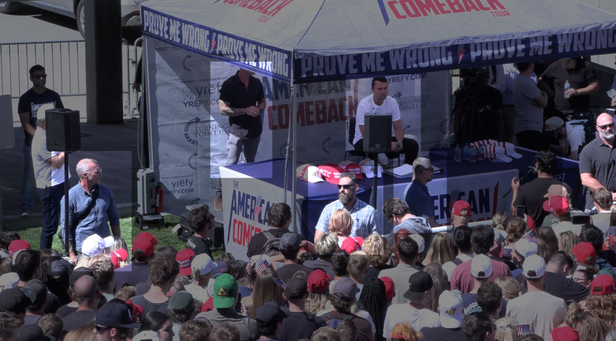 Wide view of the Turning Point USA 'American Comeback Tour' stage setup at Utah Valley University, showing Charlie Kirk seated under a tent with a microphone, staff nearby, and a large seated crowd